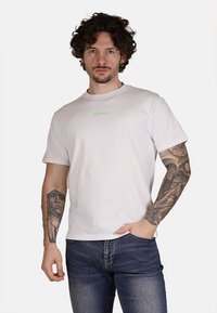 T-shirt bianco in cotone con collo alla marinara e maniche corte, caratterizzato da un piccolo testo verde "PROPAGANDA" al centro. Vestibilità casual, texture morbida.
