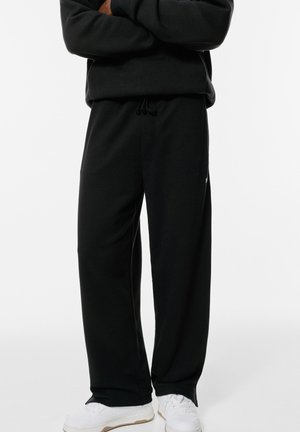 Zwarte joggingbroek van zachte stof, met een relaxte pasvorm, een trekkoord in de taille en zijzakken. Gepaard met witte sportschoenen.