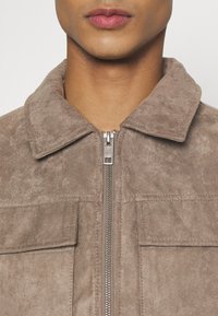 Giacca in camoscio marrone chiaro con chiusura con zip, collo con rever e due tasche frontali. Texture morbida e design pulito, adatta per un abbigliamento casual.