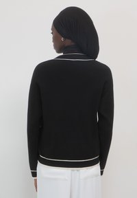 Schwarzer Pullover mit einer taillierten Passform, ausgestattet mit einem weißen Besatz entlang des Kragens und des Saums. Das Material wirkt weich und hat eine glatte Textur.