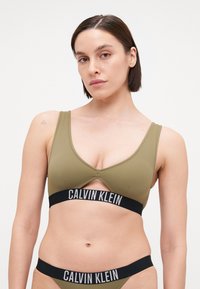 Olijf groene bralette met uitsnijdetails en brede zwarte elastische band met de "CALVIN KLEIN"-logo. Glad, rekbaar materiaal, minimale bedekking.