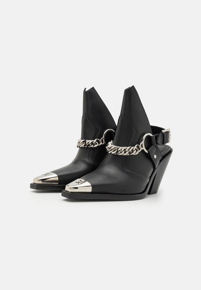 Black leather heeled shoes with pointed toe and metallic tip. Features a chain accent and adjustable buckle strap. Wedge heel design.
