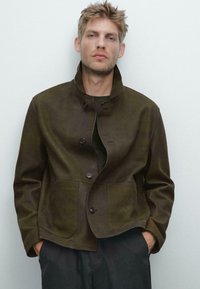 Braune Wildlederjacke mit Stehkragen, vorderem Knopfverschluss und zwei großen Taschen. Glatte Textur, lockerer Schnitt und minimale Details.