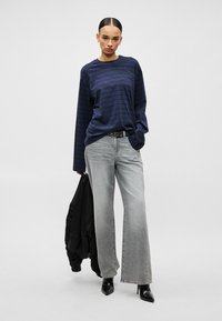 Moteris dėvi tamsiai mėlyną dryžuotą ilgomis rankovėmis marškinius, šviesiai pilkas plataus kirpimo džinsines kelnes, juodus aštriais galiukais aukštakulnius, vienoje rankoje laiko juodą striuk�ę.