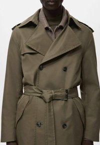 Abrigo tipo trench verde oliva con diseño de doble botonadura, cuello ancho y cintura con cinturón; cuenta con hombreras y bolsillos laterales.