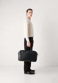 Svart lær laptopbag med to håndtak, glidelås sidelomme, glatt tekstur og avrundet form. Båret av en modell i en lys genser.