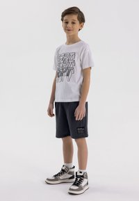 Witte t-shirt met korte mouwen met zwarte grafische tekst, gecombineerd met marineblauwe shorts en grijze hoge sneakers met zwart-witte accenten.