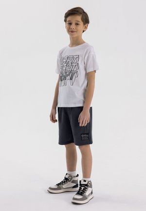 T-shirt bianco a maniche corte con testo grafico nero, abbinato a pantaloni corti blu navy e sneaker alte grigie con dettagli in bianco e nero.