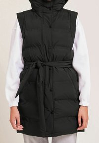 Czarna pikowana kamizelka bez rękawów z kapturem, z paskiem w talii, zatrzaskami i bocznymi kieszeniami; w zestawie z białą koszulą z długim rękawem.