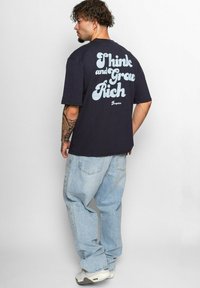 Navyblå bomulds t-shirt med kontrastfyldt lyseblå tekst på ryggen, der siger "Think and Grow Rich." Løs pasform kombineret med lyse denim jeans.
