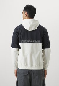 Giacca a vento bianca e nera con cappuccio, dotata di polsini elasticizzati, orlo arricciato e logo 'UNDER ARMOUR' sulla schiena.