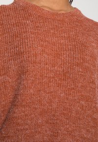 Pull en maille de couleur terre cuite chaude, avec un tissage texturé, un col rond et un tissu doux, présentant des fibres visibles et une légère variation de ton.