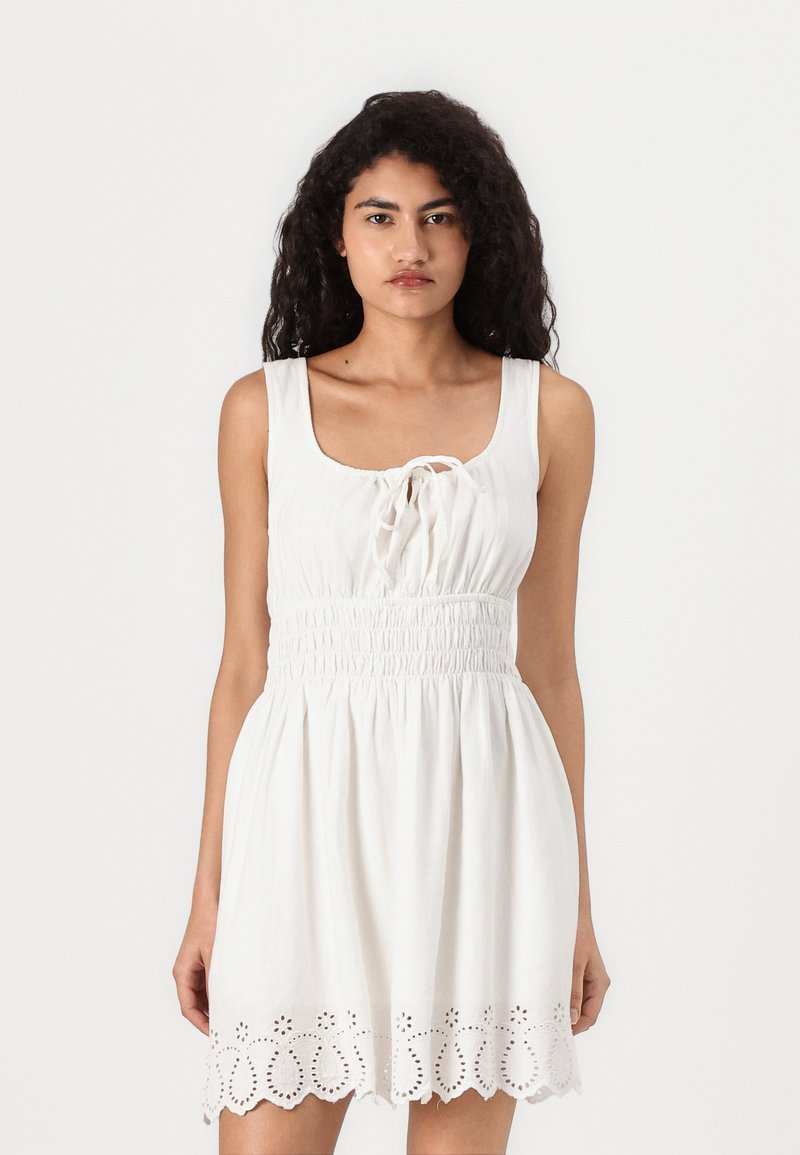 White dress with a scoop neckline, adjustable tie at the front, elastic smocked waistband, and a decorative eyelet hem.
