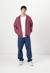 Hanorac zip-up în nuanță maroniu, tricou cu guler rotund alb, blugi largi albaștri cu detalii din materiale patch, și adidași albi. Fundal simplu.