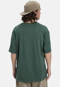 Jonge persoon met lang krullend haar, een beige pet achterstevoren, een donkergroene oversized t-shirt en lichtbruine broek, zich van de camera afkerend.