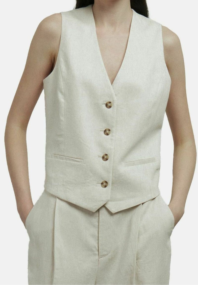 Femme portant un gilet beige sans manches boutonné et un pantalon taille haute assorti, les mains dans les poches, sur un fond uni.