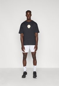 Must mustaheline T-särk kuldse Nike logo ja valgete lühikeste pükste ning mustade spordijalatsitega, seismas lihtsa tausta ees.