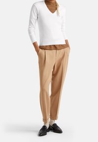 Biały sweter na V z dzianiny, nałożony na brązowy top, zestawiony z beżowymi plisowanymi spodniami i czarnymi mokasynami z wzorzystymi skarpetkami.