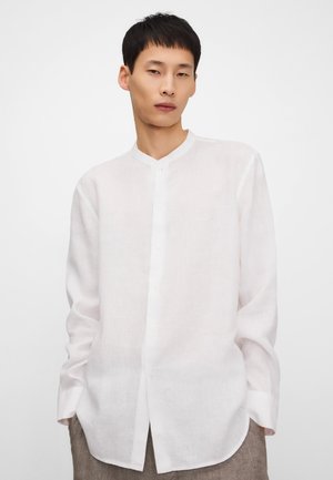 Junger Mann trägt ein weißes, langärmliges Hemd mit Knopfleiste und Stehkragen, steht vor einem schlichten hellen Hintergrund.