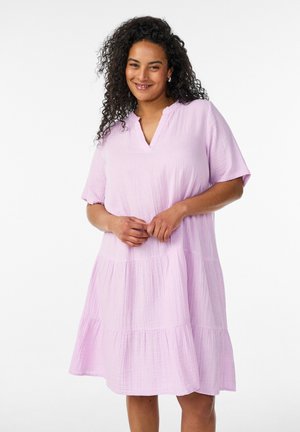 Usmievavá žena s kučeravými vlasmi nosiaca voľné, svetlofialové, krátke rukávy, kolenné šaty s vrstvenou sukňou, stojaca na bielom pozadí.