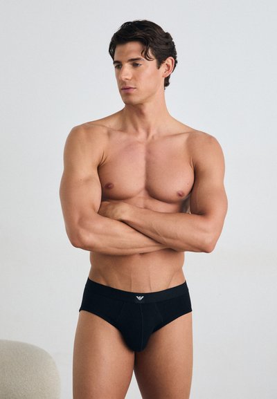 Zwarte slips van zacht stof, met een gevormde pouch en een gladde tailleband met logo. Model heeft een atletisch figuur en heeft de armen gekruist.