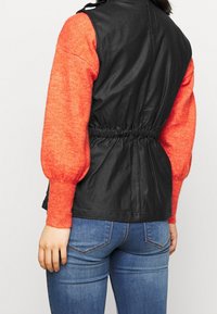 Svart läderväst med en figursydd midja, ihop med en ljusorange, tjockstickad tröja. Blåjeans fulländar den avslappnade outfiten.