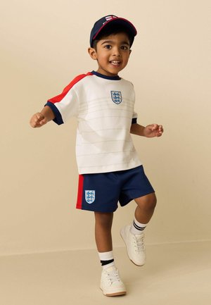 Junger Junge, der den Fußballtrikot von England mit weißem Shirt, blauen Shorts, Kappe, weißen Turnschuhen und gestreiften Socken trägt, posiert mit einem angehobenen Bein.
