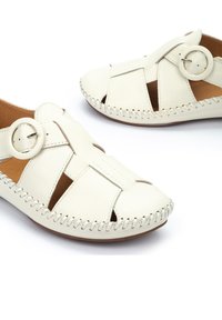 White leather shoes with cut-out design, round buckle embellishment, and contrasting stitching along the edge.