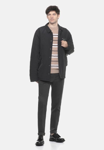 Jonge man staat, gekleed in een zwarte jas, beige gestreepte trui, zwarte broek en zwarte nette schoenen, terwijl hij een zwart rugzakriem vasthoudt.