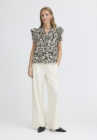 Blusa a fiori nera e crema con maniche a ruffle, abbinata a pantaloni bianchi a gamba larga e scarpe nere a punta.