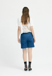Denim lühikesed püksid franseeritud servaga, koos valge graafilise T-särgiga, millel on kalamuster. Mustad sokid ja kontsaga sandaalid täiendavad riietust.