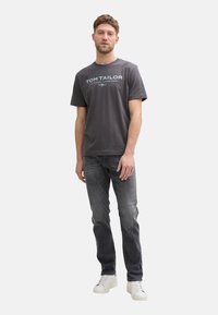 Tricou din bumbac gri cu text turcoaz "TOM TAILOR", guler rotund, mâneci scurte. Asortat cu blugi închisi și adidași albi.