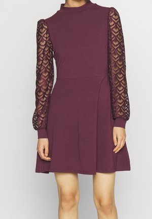 Krótka sukienka damska w kolorze śliwkowym z wysokim kołnierzem i długimi, przejrzystymi koronkowymi rękawami z geometrycznymi wzorami.