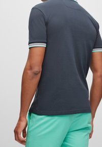 Man som bär en mörkgrå kortärmad polotröja med randiga ärmslut och mintgröna shorts, står mot en neutral bakgrund.