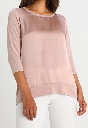Femme portant un haut transparent rose clair à manches longues avec un col rond et un pantalon blanc.