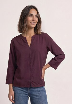 Femme aux cheveux foncés mi-longs portant un chemisier bordeaux boutonné et un jean bleu, souriante et regardant sur le côté avec une main sur la hanche.