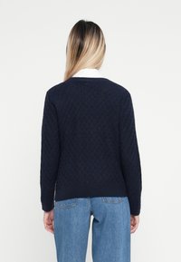 Suéter de malha azul marinho com padrão de diamante, gola redonda e bainha canelada, usado sobre jeans de ganga, mostrando um design de tecido texturizado.