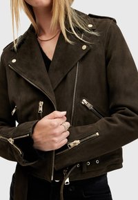 Giacca biker in finta suede verde scuro con zip, bottoni a scatto e un design leggermente corto. Presenta tasche laterali con chiusura a zip.
