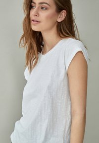 Camiseta blanca de manga corta en una tela ligera y texturizada con un corte relajado y cuello redondo, que presenta un diseño sencillo.
