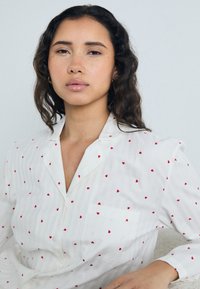Camicia bianca con bottoni, decorata con piccoli motivi a forma di cuore rossi, dotata di taschino, colletto e maniche lunghe con una finitura leggermente texturizzata.