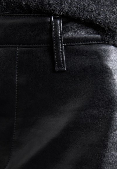Pantaloni in pelle nera con una texture liscia, caratterizzati da una vita cucita e una chiusura a gancio metallico. La vista ravvicinata mette in evidenza i dettagli del materiale.