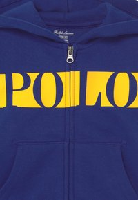 Modrá mikina s kapucí na zip s velkým žlutým grafikou "POLO". Vyrobena z bavlněné směsi, má vpředu kapsu a kovový zip.