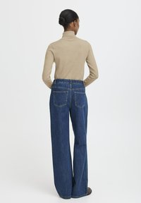 Donna con i capelli raccolti in un chignon, che indossa un dolcevita beige e jeans a vita alta larghi di colore blu, in piedi di spalle su uno sfondo neutro.