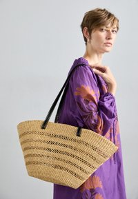 Kudottu olkikassi avoimella kudonnalla, mustat nahkahihnat ja kulmikas muoto, yhdistettynä violettiin brodeerattuun mekkoon.