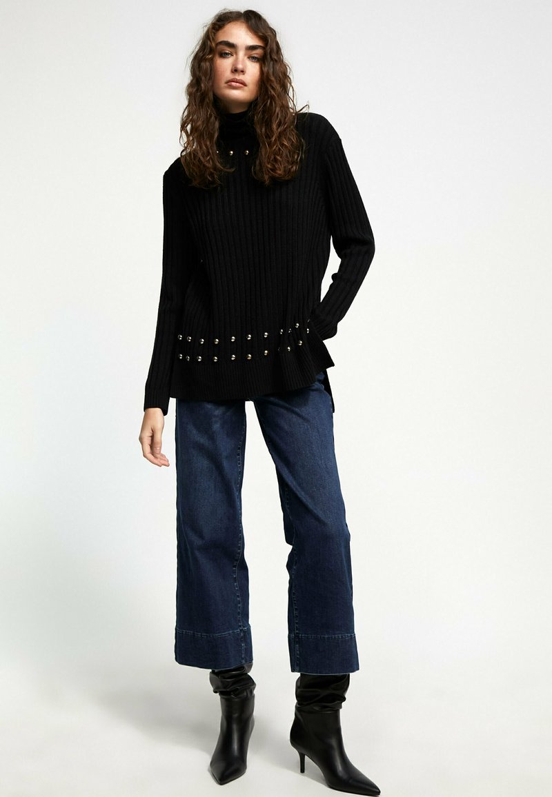 Black ribbed turtleneck sweater with metallic studs at the hem, paired with wide-leg denim jeans and black pointed ankle boots.
