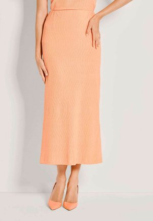 Vrouw die een perzikkleurige midirok en bijpassende puntige hoge hakken draagt, staand tegen een effen witte achtergrond.