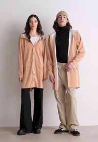 Two individuals wear beige rain jackets with white accents, featuring snap buttons and pockets. They are paired with wide-leg trousers.