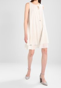Ärmelloses weißes knielanges Kleid mit dezentem Stickmuster, mit Krawatte am Hals, getragen mit grauen Blockabsatzschuhen mit Knöchelriemchen vor hellem Hintergrund.