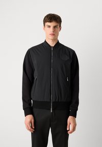 Čierna bomber bunda s hladkým, lesklým materiálom, rebrovanými manžetami a zipsom. Obsahuje textúrovaný emblém na hrudi.