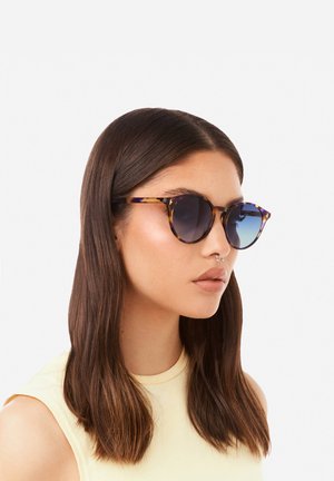 Ochelari de soare tortoise cu lentile gradiente albastre, formă rotundă și brațe subțiri. Modelul poartă un top galben și are păr lung și drept.